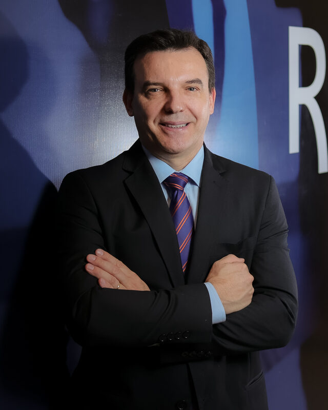 Edimar José Niec, sócio-fundador da Recon, de terno escuro e braços cruzados, diante de painel com a marca da empresa